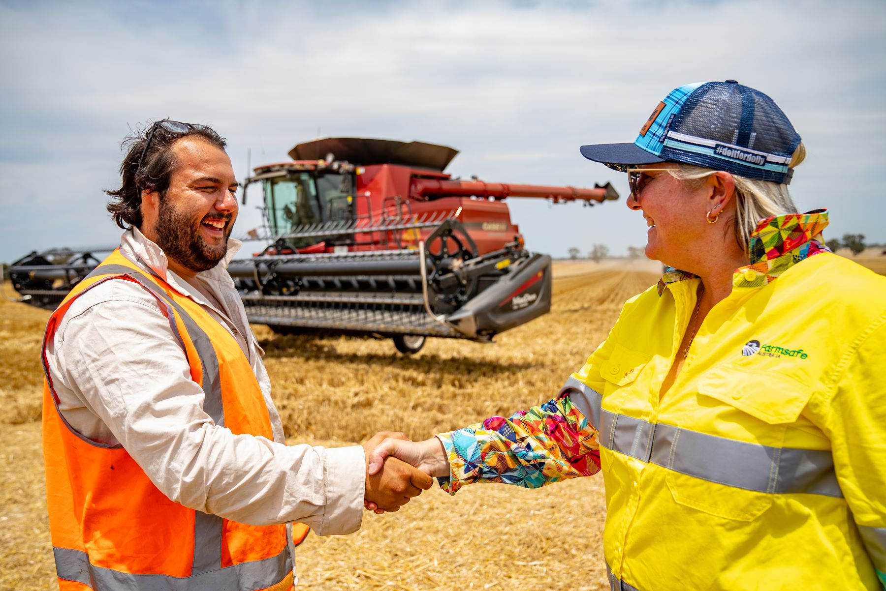 Man and woman shaking hand in paddock on farm with header behind them.